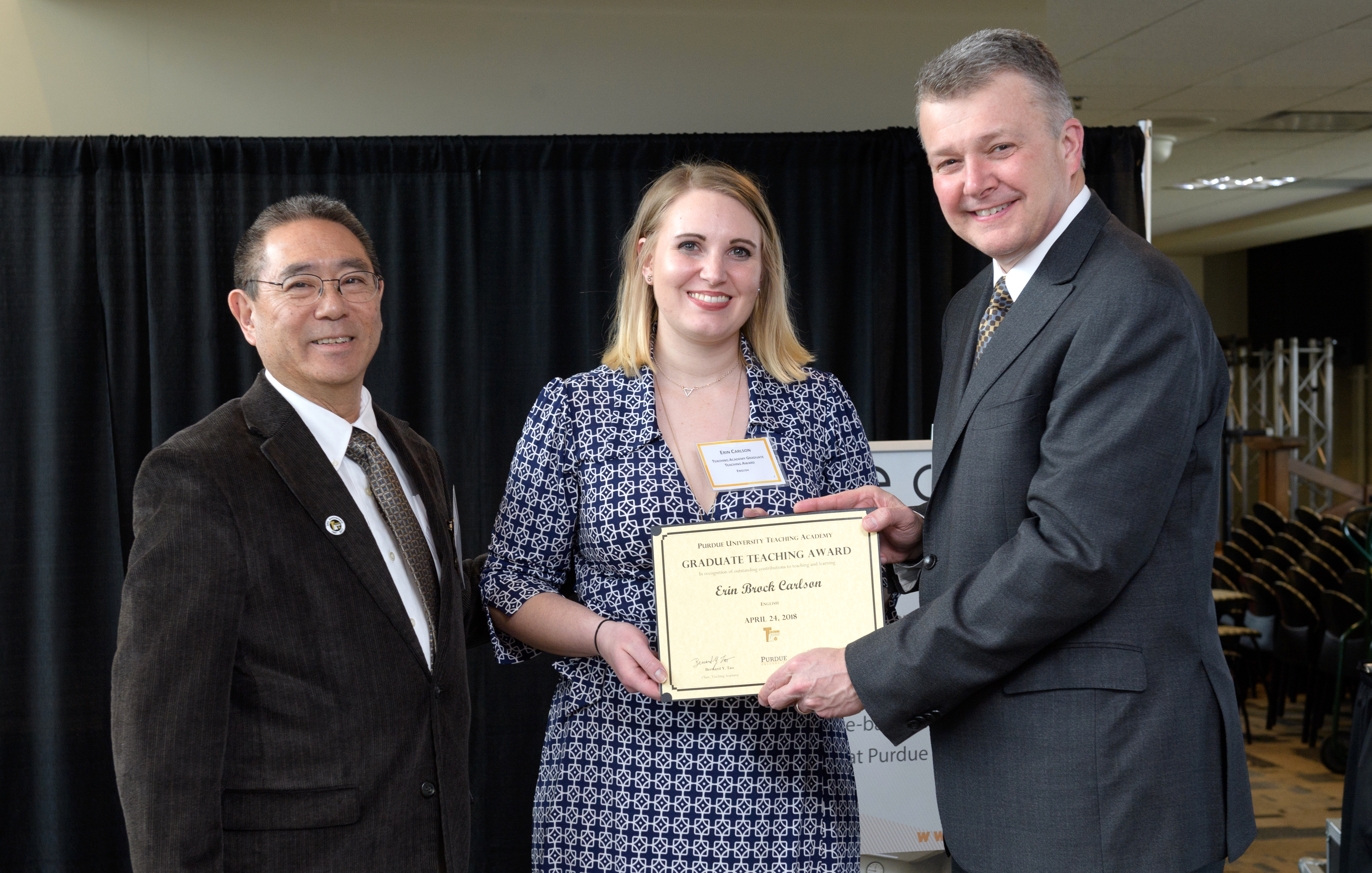 Blonde woman smiling and holding certificate in between two older men with suits.